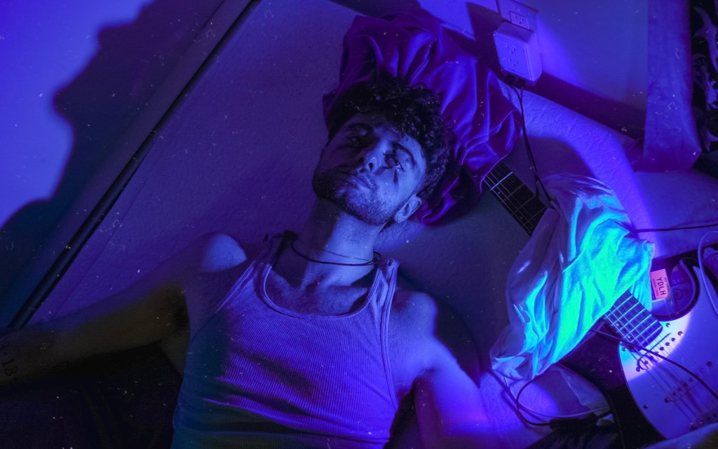 Scxtty lying on the floor with a guitar and makeup on his face. A blue light covers the room.