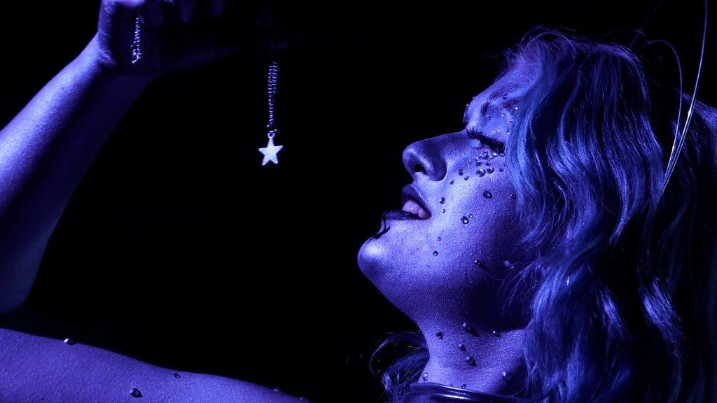 Kally Hall stands illuminated by blue light while holding a necklace with a star in front of her face.