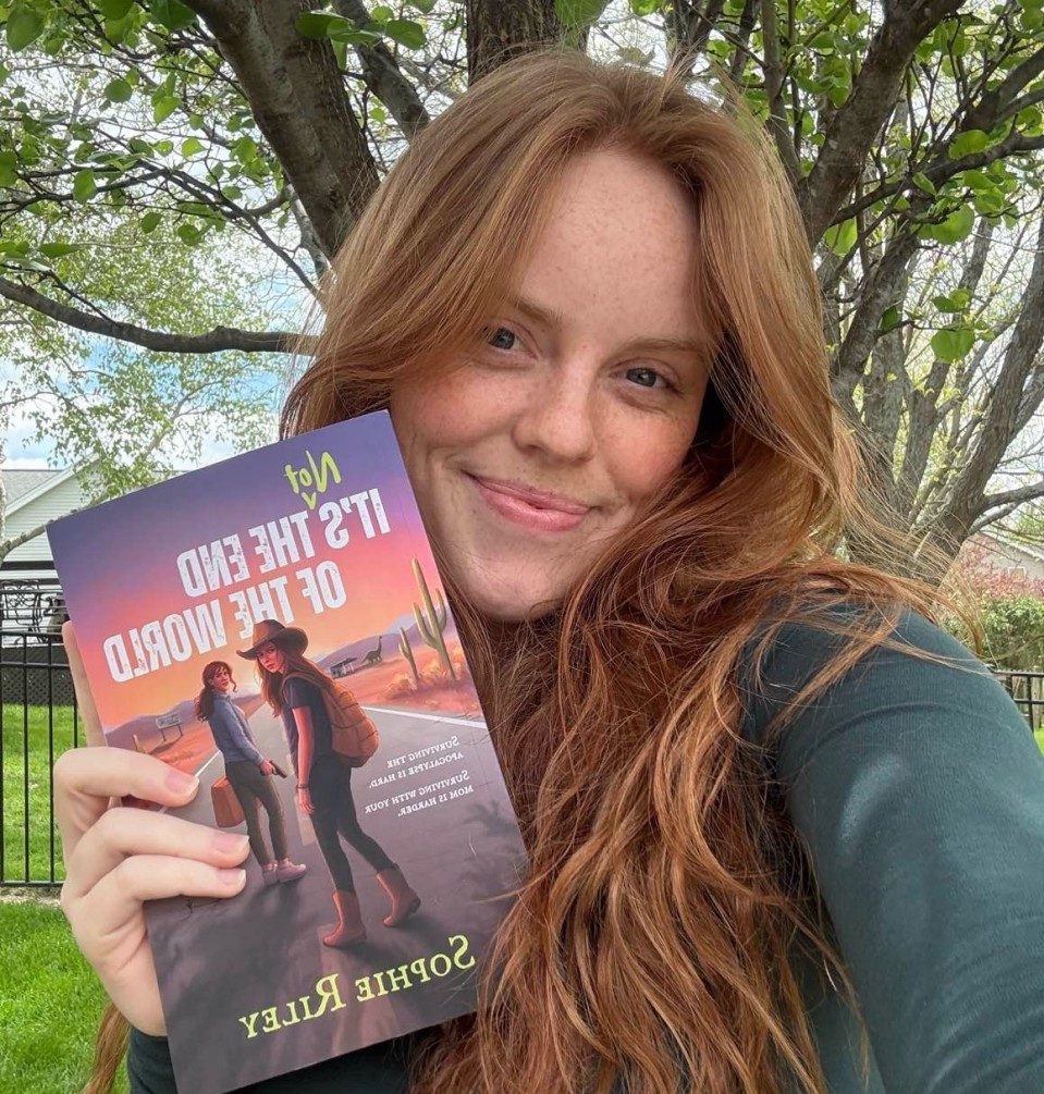 Sophie Riley, a white woman with blonde-red hair, poses in a green long-sleeve T-shirt with her debut novel, It's Not the End of the World, with trees in the background.