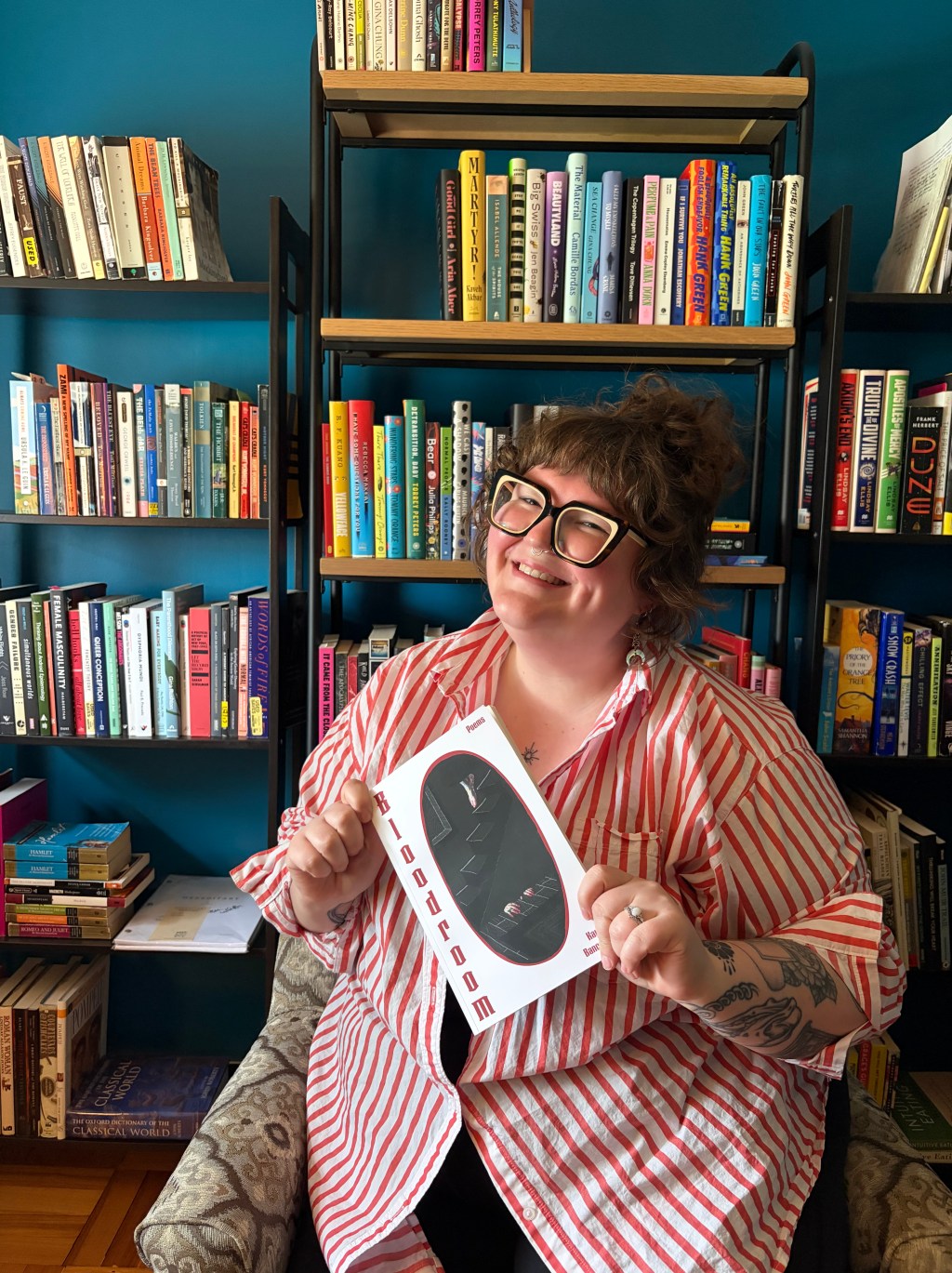 Kay E. Bancroft sitting and holding a copy of their debut poetry collection, "Bloodroom."