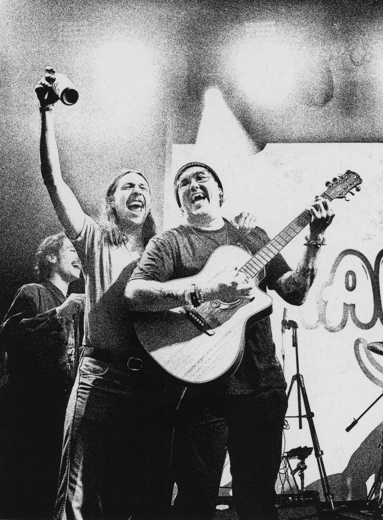 Man playing guitar and man holding beer smile onstage at concert